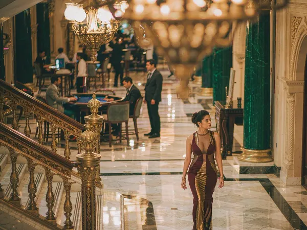 playrupees Woman in a maroon and gold dress walking through a chandelier-lit casino lobby